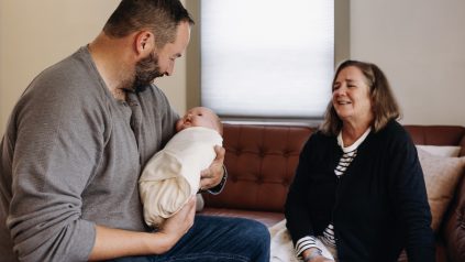 Man sitting on a couch holding a baby doll wrapped in a blanket, woman sitting nearby watching